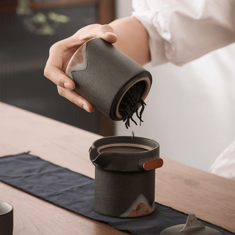 Person pouring from a ceramic container into another on a wooden table.