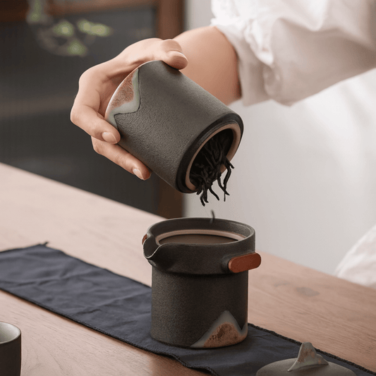 Person pouring from a ceramic container into another on a wooden table.