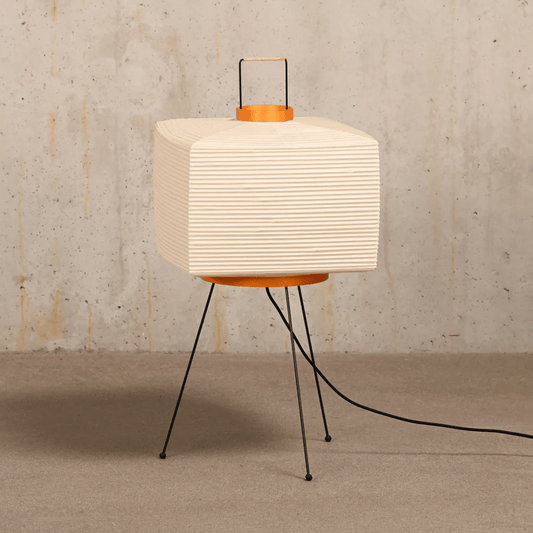 Table lamp with a white paper-like shade and black metal legs against a textured wall.