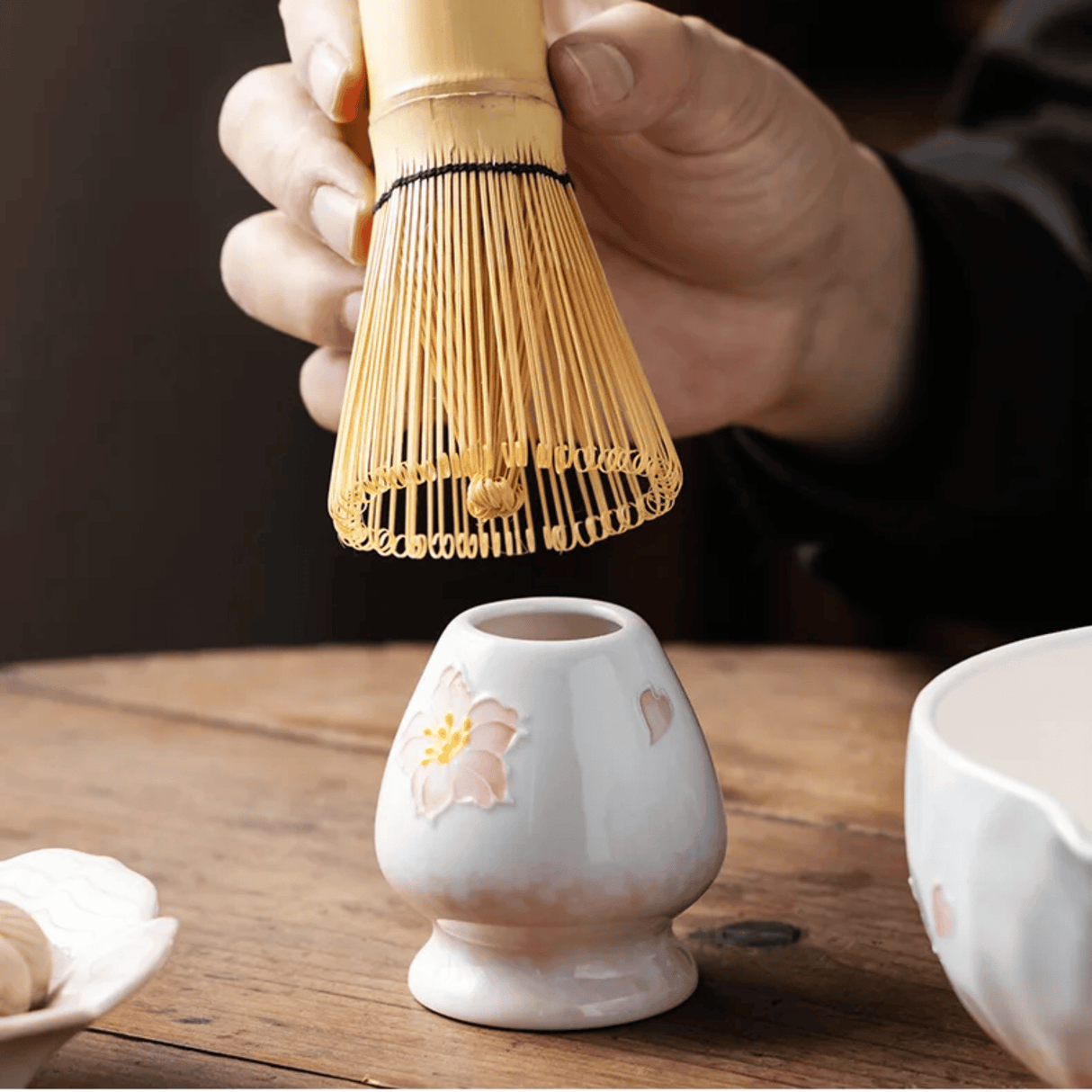 Person using a bamboo whisk over a small ceramic container on a wooden surface