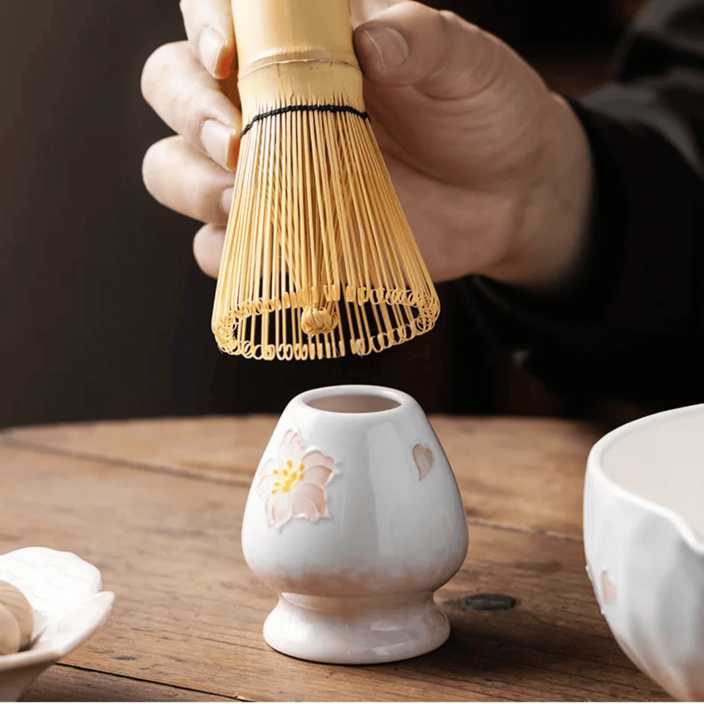 Person using a bamboo whisk over a small ceramic container on a wooden surface