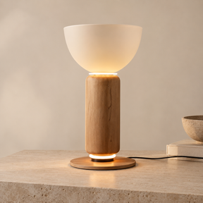 A modern table lamp with a frosted white bowl-shaped shade, a cylindrical natural wood base, and a round wooden footrest, shown lit on a stone tabletop against a warm beige wall.