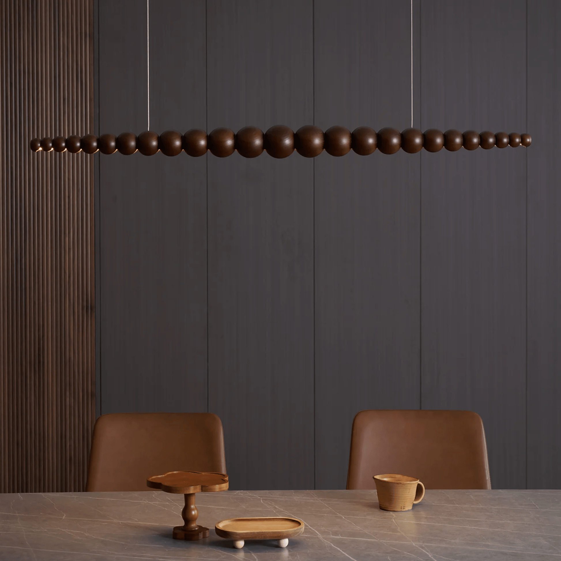 Wooden pendant light fixture above a dining table with chairs against a dark paneled wall.