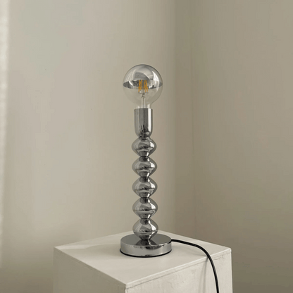 Silver table lamp with a unique design on a white surface against a beige wall.