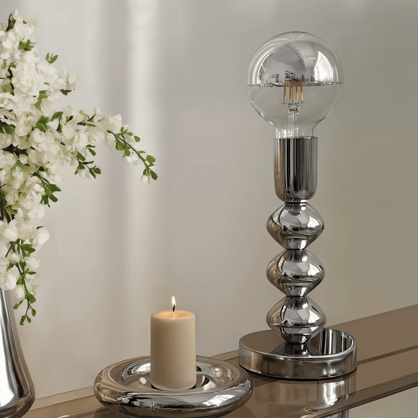 Silver table lamp with a candle on a reflective surface, with white flowers in the background.