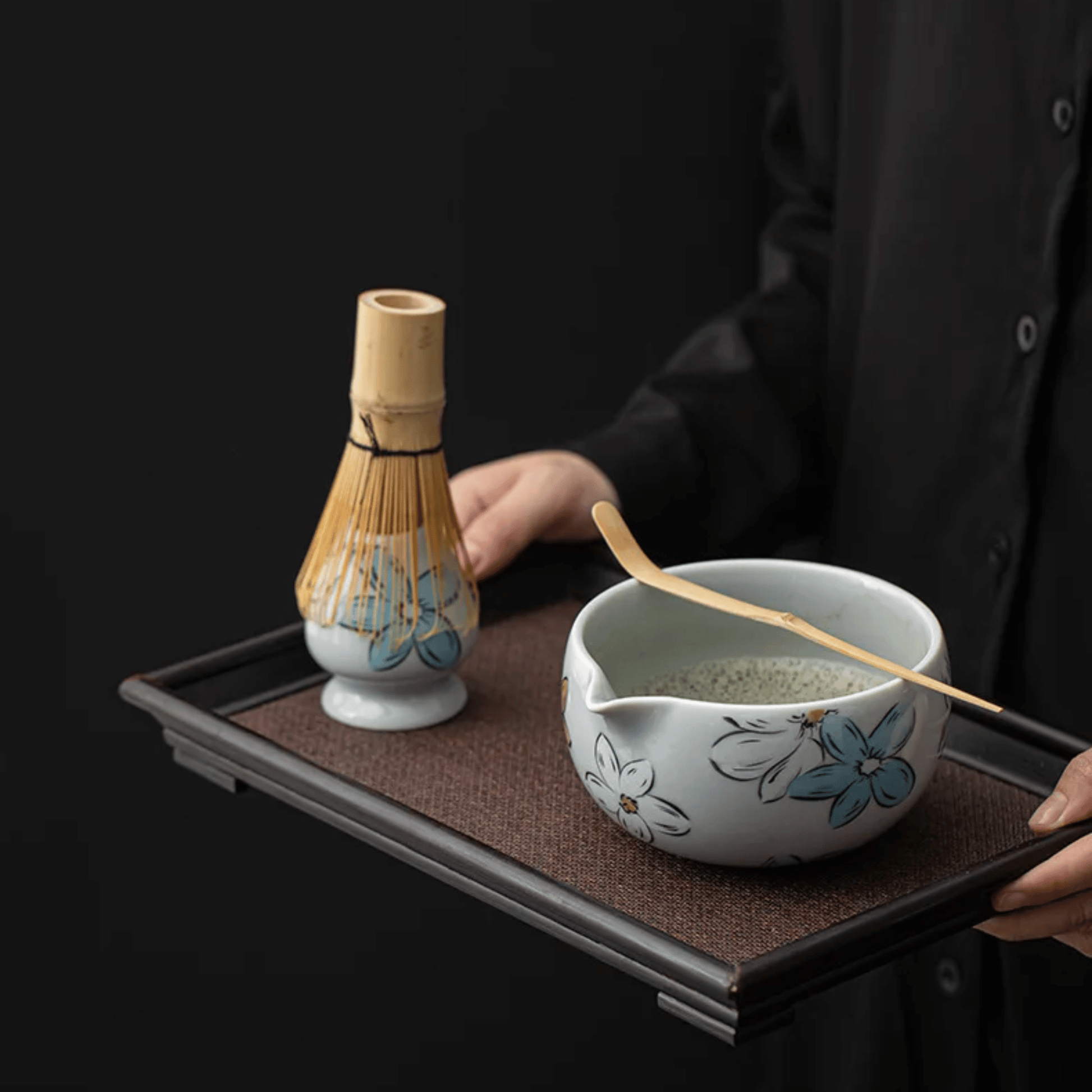 Person holding a tray with a ceramic bowl and whisk, against a black background