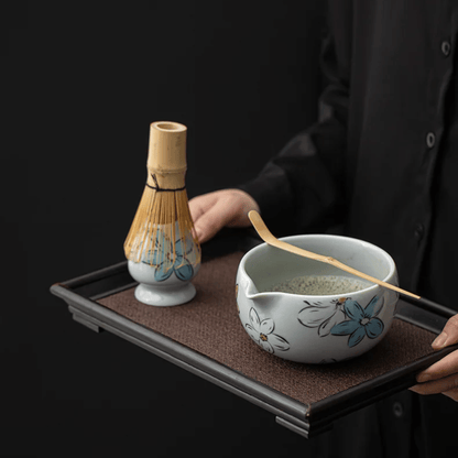 Person holding a tray with a ceramic bowl and whisk, against a black background