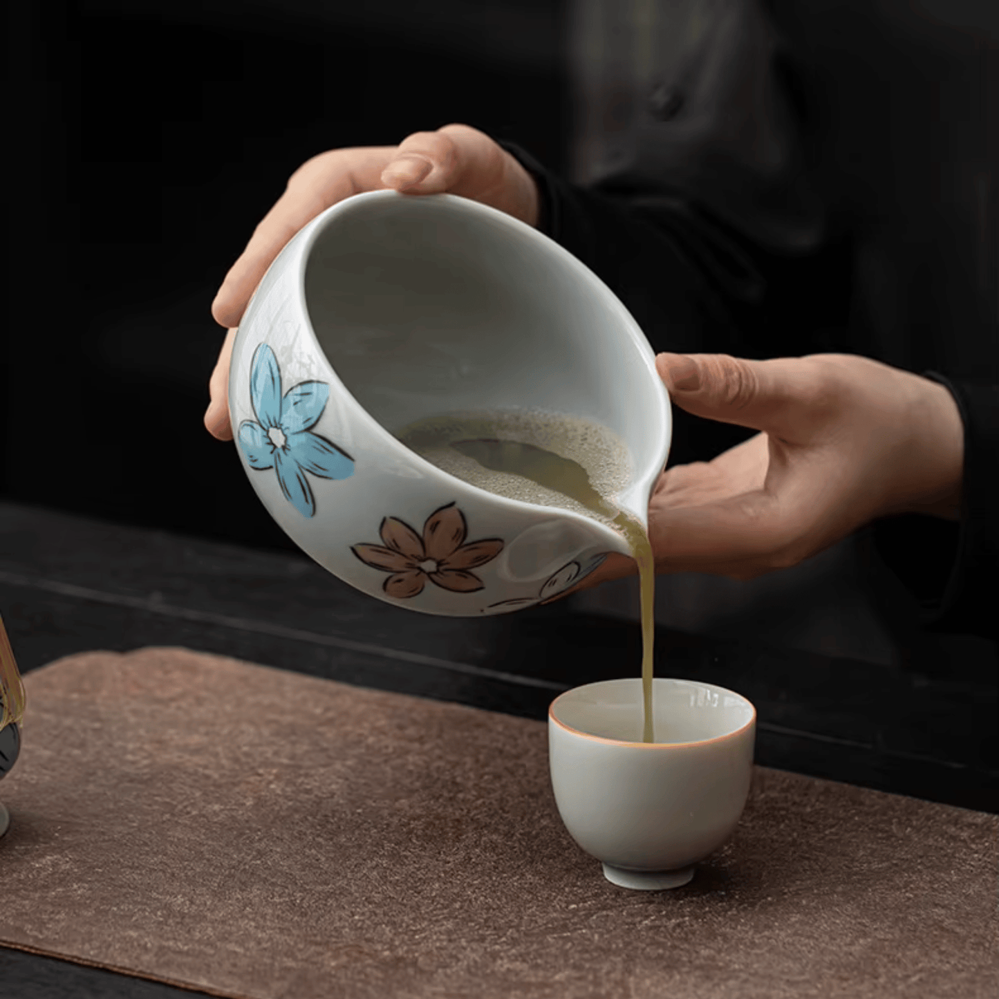 Person pouring tea from a decorative ceramic cup into another cup on a dark surface.