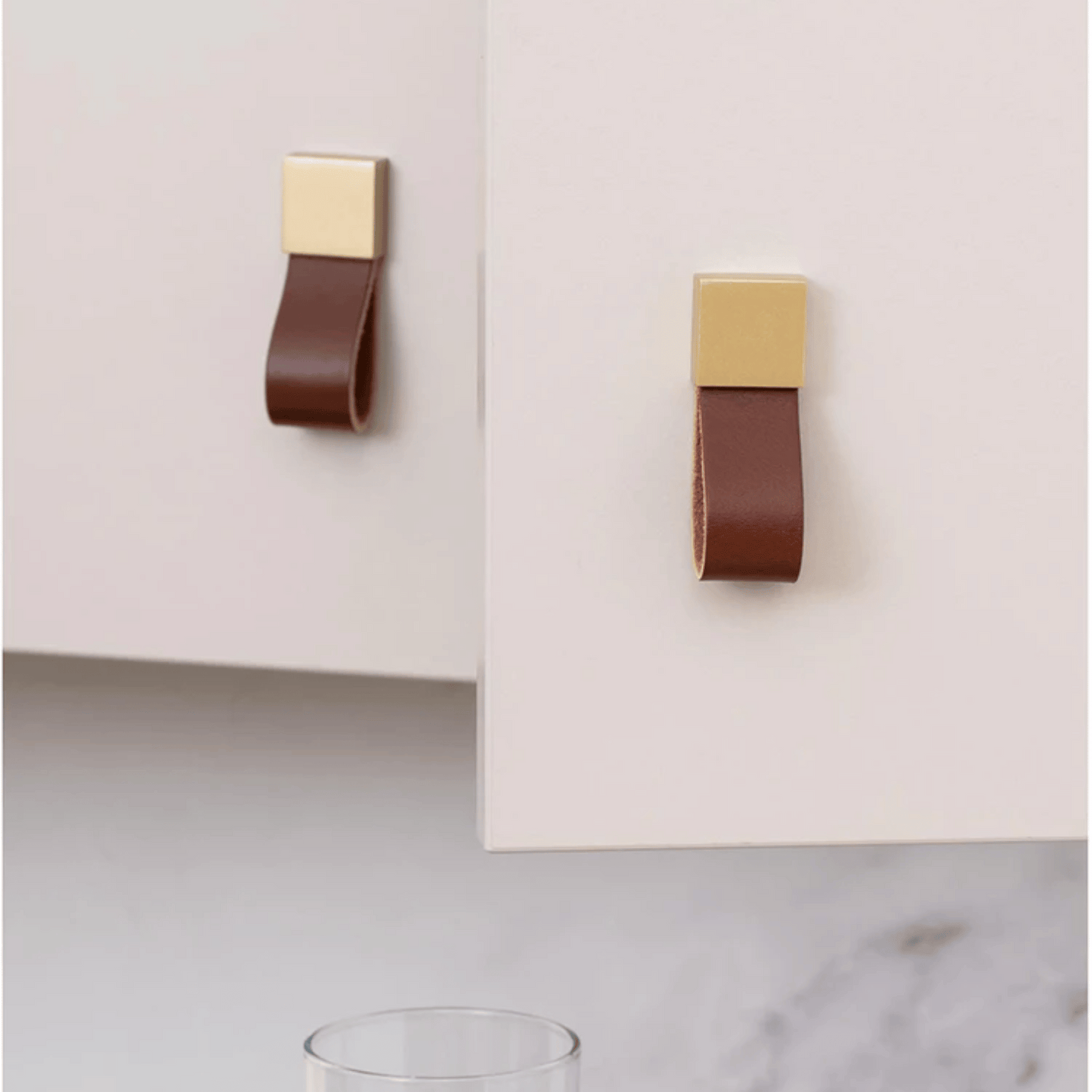 Two brown leather handles with gold metal accents on a white cabinet.