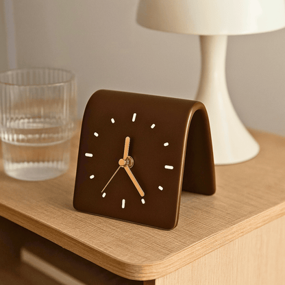 Brown square clock on a wooden surface with a glass and lamp in the background