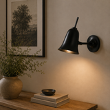 Black wall lamp on a wall with a vase and books on a wooden surface below.