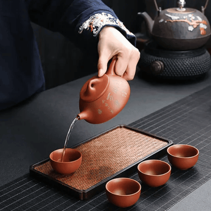 Person pouring tea from a ceramic teapot into cups on a dark surface with a teapot in the background.