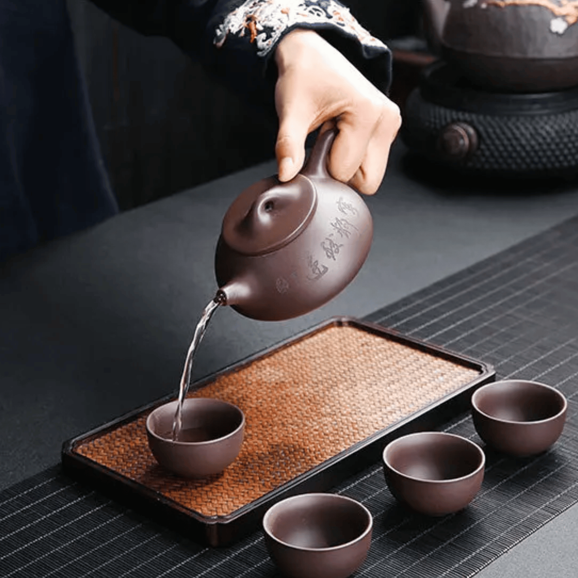 Person pouring tea from a ceramic teapot into cups on a dark surface.