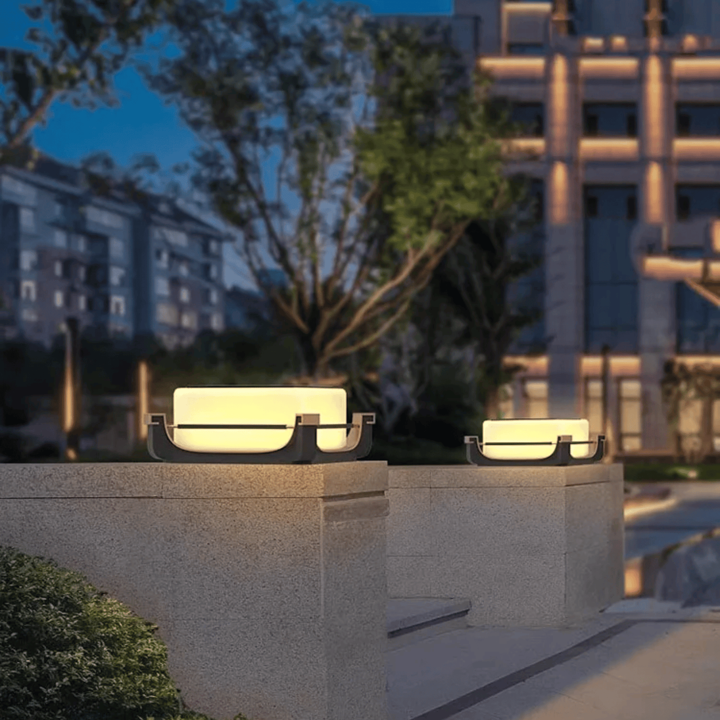 Two outdoor lights on stone plinths with a blurred building and tree in the background.