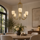 A modern brass chandelier with five frosted glass shades hangs over a dining table in a beautifully styled dining room with beige walls, a large window, and a vase of white flowers.