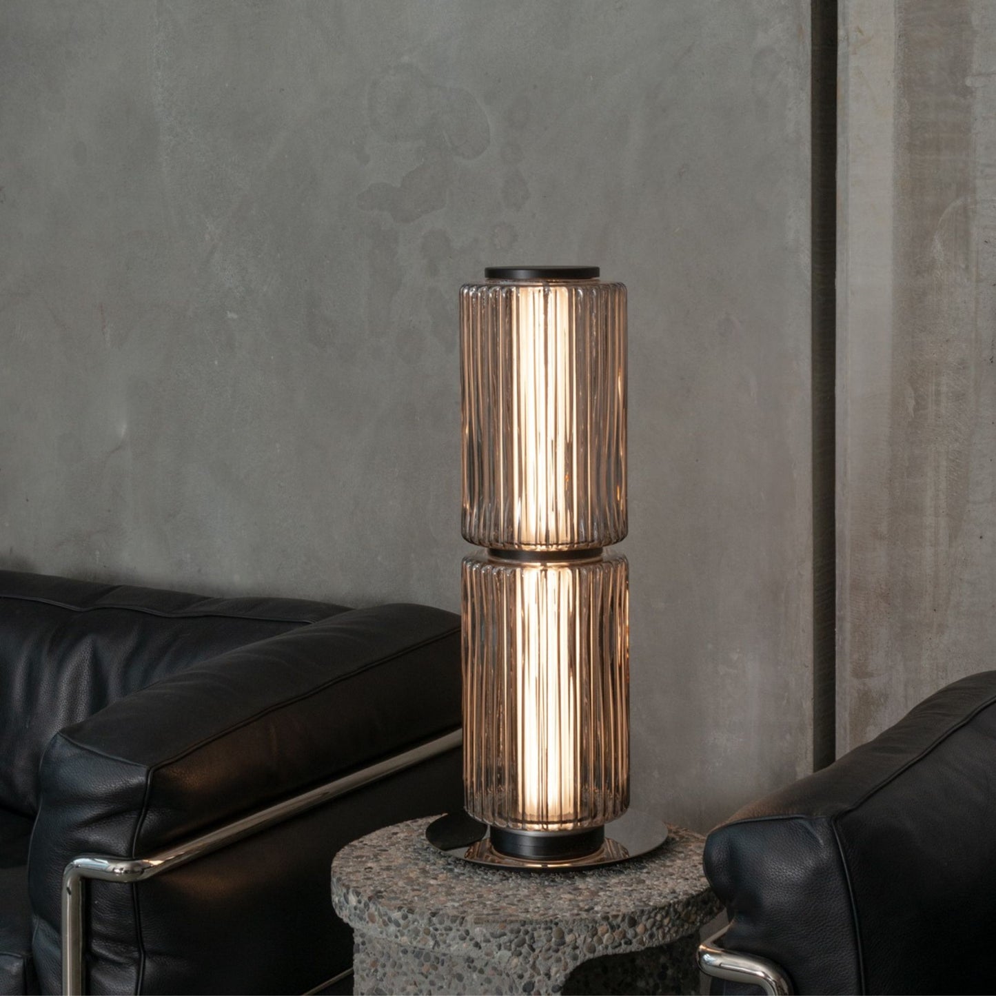 Double-stacked ribbed-glass lamp emitting warm light on a modern glass table near a gray sofa.