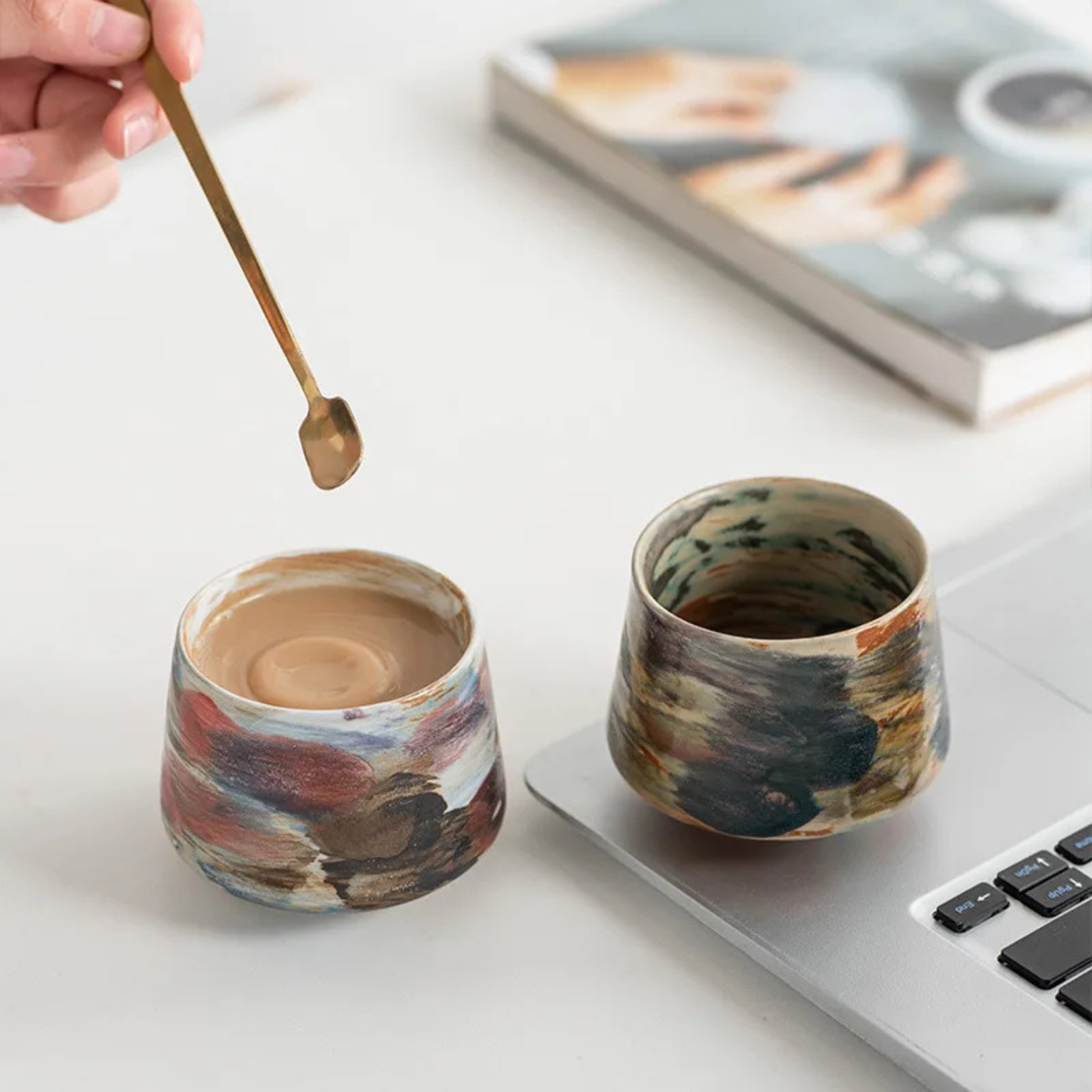 Two artisan ceramic cups with abstract multicolor glazes being held in both hands for display.