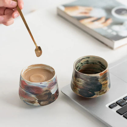 Two artisan ceramic cups with abstract multicolor glazes being held in both hands for display.