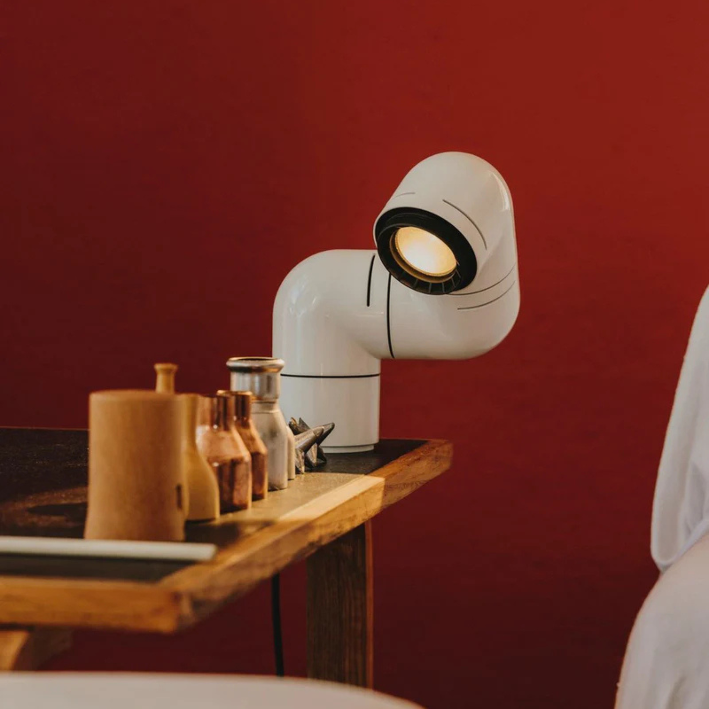 A white modern desk lamp in the shape of an elbow pipe, casting light onto a wooden table with various objects.