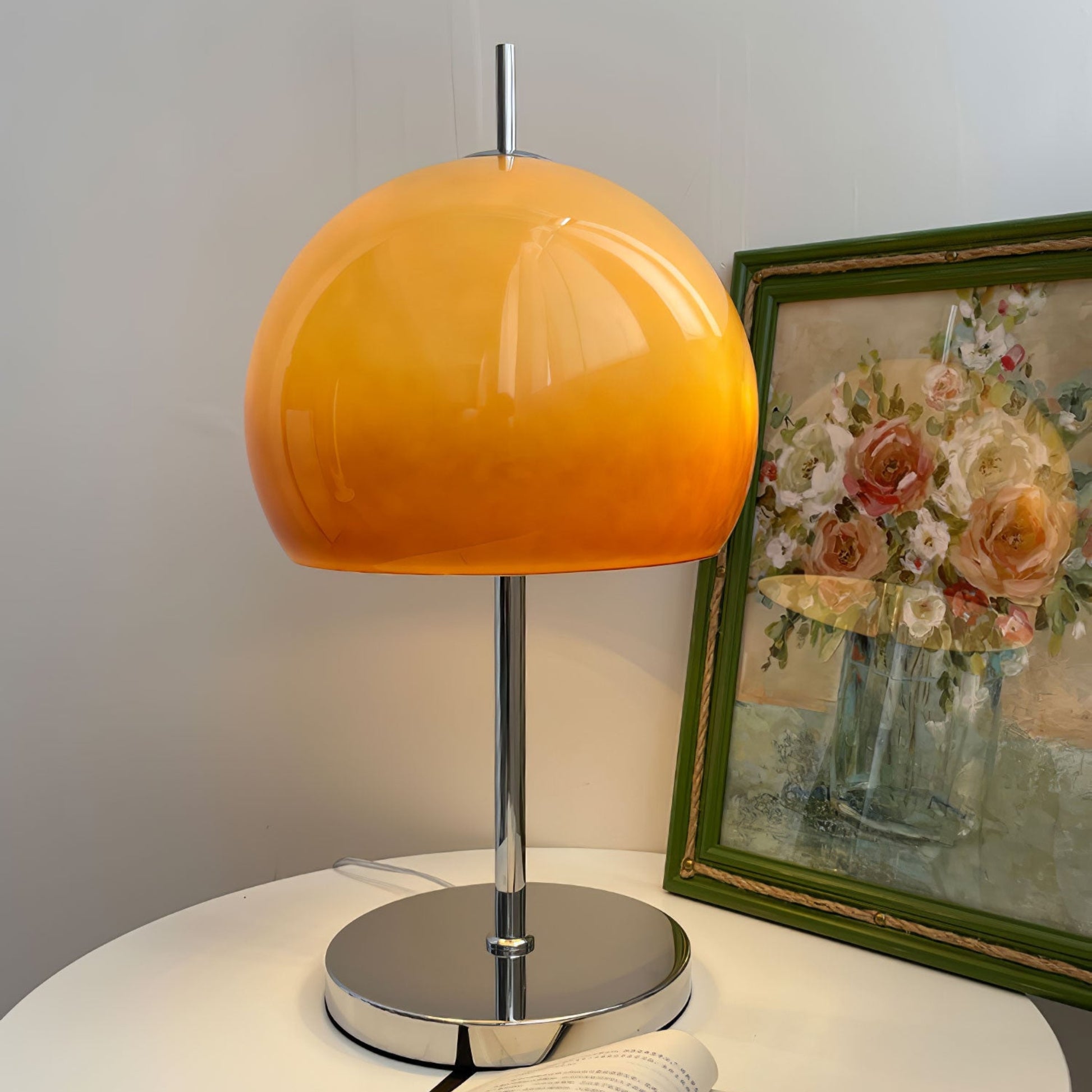 A sleek table lamp with a orange globe shade placed on a simple desk next to an open window.