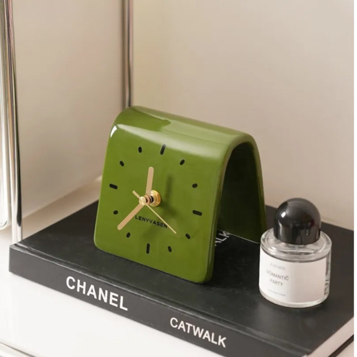 A green leather desk clock with gold hands and black hour markers placed on a “CHANEL CATWALK” book beside a small round perfume bottle.