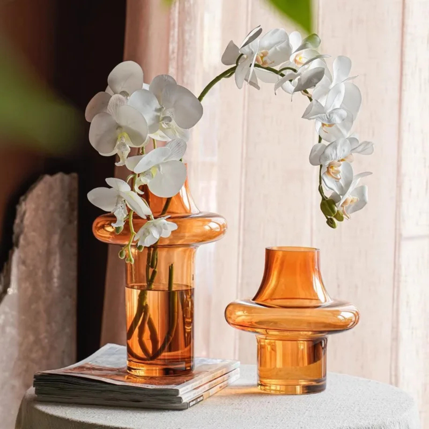 Two amber glass vases on a table near a window, each holding cascading white orchid flowers, with soft natural light.