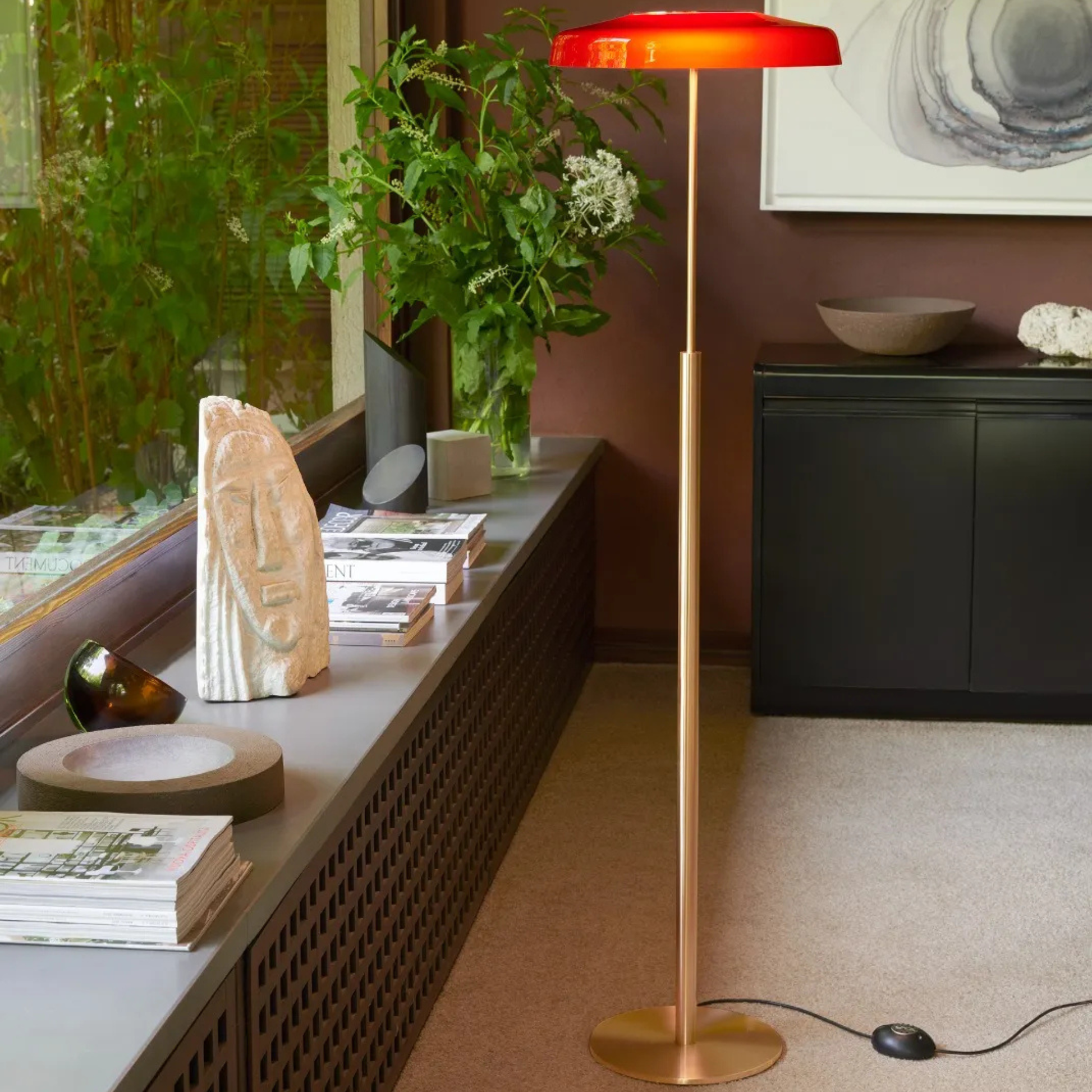 A modern floor lamp with a red circular shade standing beside a window and green plants in a cozy, well-decorated living room.