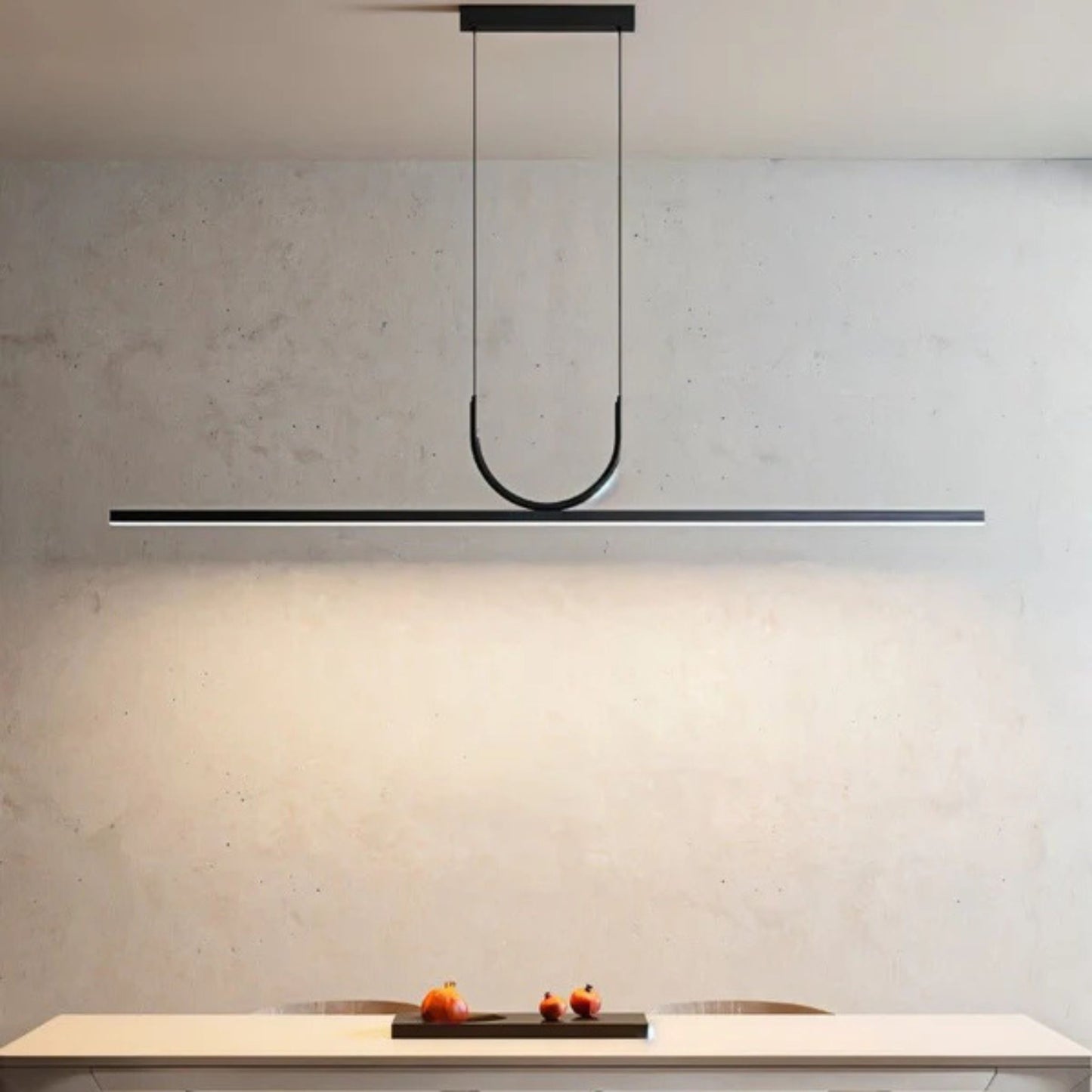 A minimalist black pendant light with a linear design, casting soft light on a beige wall above a countertop.