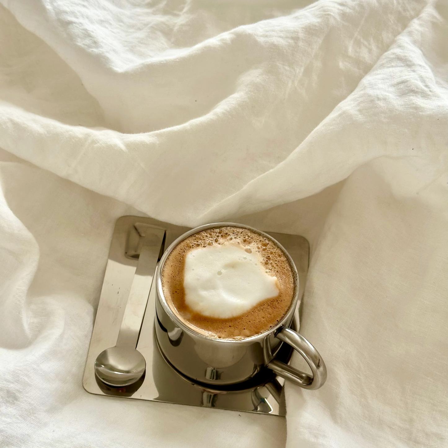 Coffee served in a stainless steel cup on a reflective saucer placed on soft white bedding.