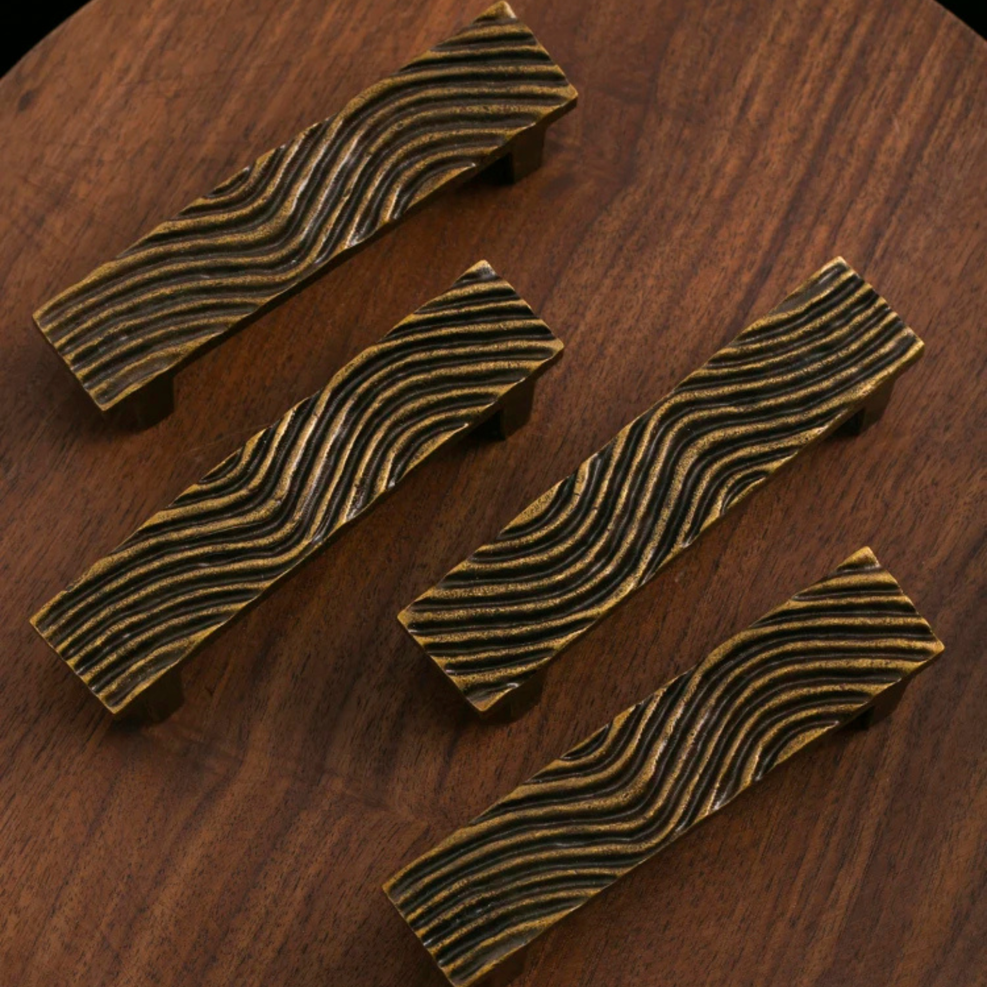 Four rectangular brass drawer handles with wavy engraved patterns placed on a wooden surface.