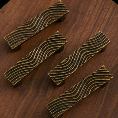 Four rectangular brass drawer handles with wavy engraved patterns placed on a wooden surface.