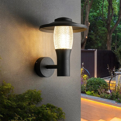 A modern black outdoor wall sconce with a textured glass shade is mounted on a gray wall, emitting warm light. In the background, there is a landscaped garden with greenery, trees, and soft pathway lighting.
