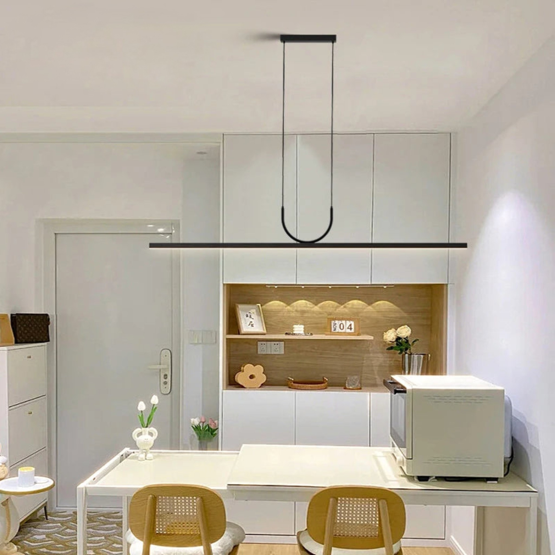 A modern, minimal kitchen with white cabinets, a built-in wooden shelf, a white microwave on the counter, two rattan chairs, and a sleek black pendant light hanging above a small dining table.