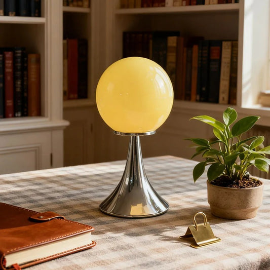 A yellow glass globe table lamp with a chrome cone-shaped base placed on a checkered tablecloth in a cozy room with bookshelves.