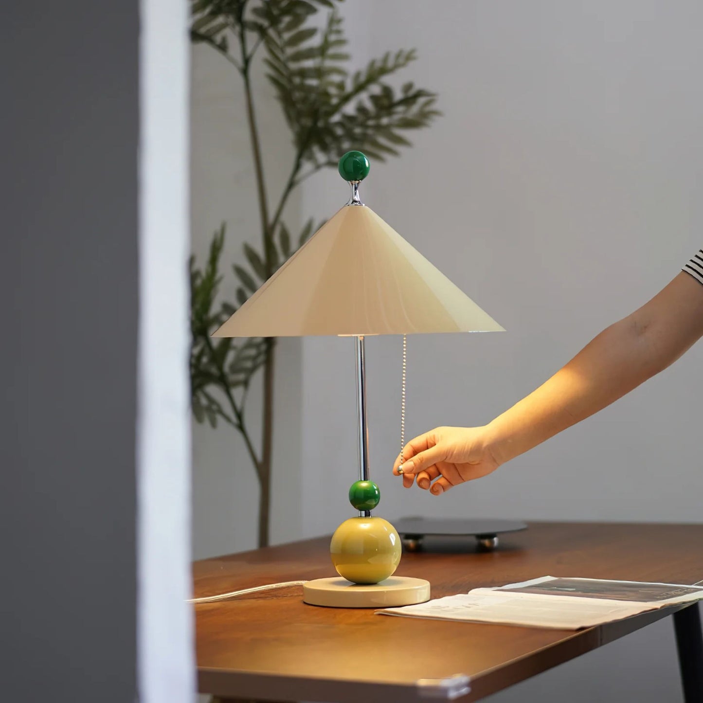 A hand adjusting the green accent ball on a beige cone-shaped table lamp on a wooden surface.