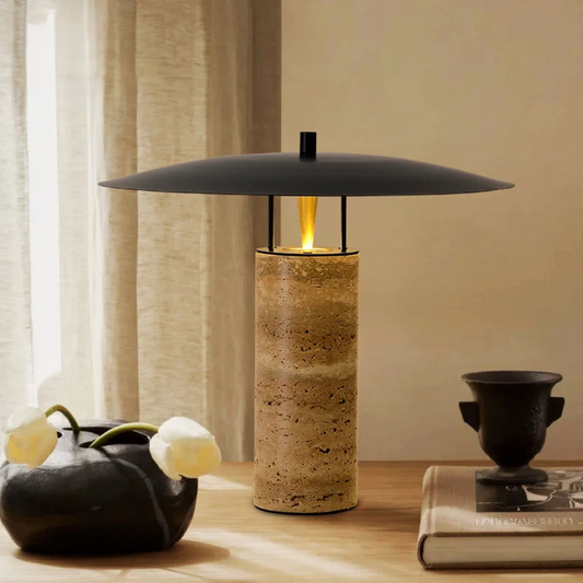 A modern table lamp with a stone cylindrical base and wide, flat black shade sits on a wooden table, next to a black vase with white flowers, a ceramic cup, and a closed book near a window with sheer curtains.