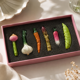 A set of six ceramic vegetable-shaped ornaments or figurines displayed in a pink gift box with black foam insert. The vegetables include a pink and green onion, a white and green garlic bulb, an orange carrot with green tops, a green and brown asparagus piece, a pink and white radish, and a green pea pod.