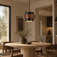 A modern cylindrical pendant light with a black top and bottom band and a warm wood-grain middle band, hanging over a round stone dining table in a neutral-toned dining room.