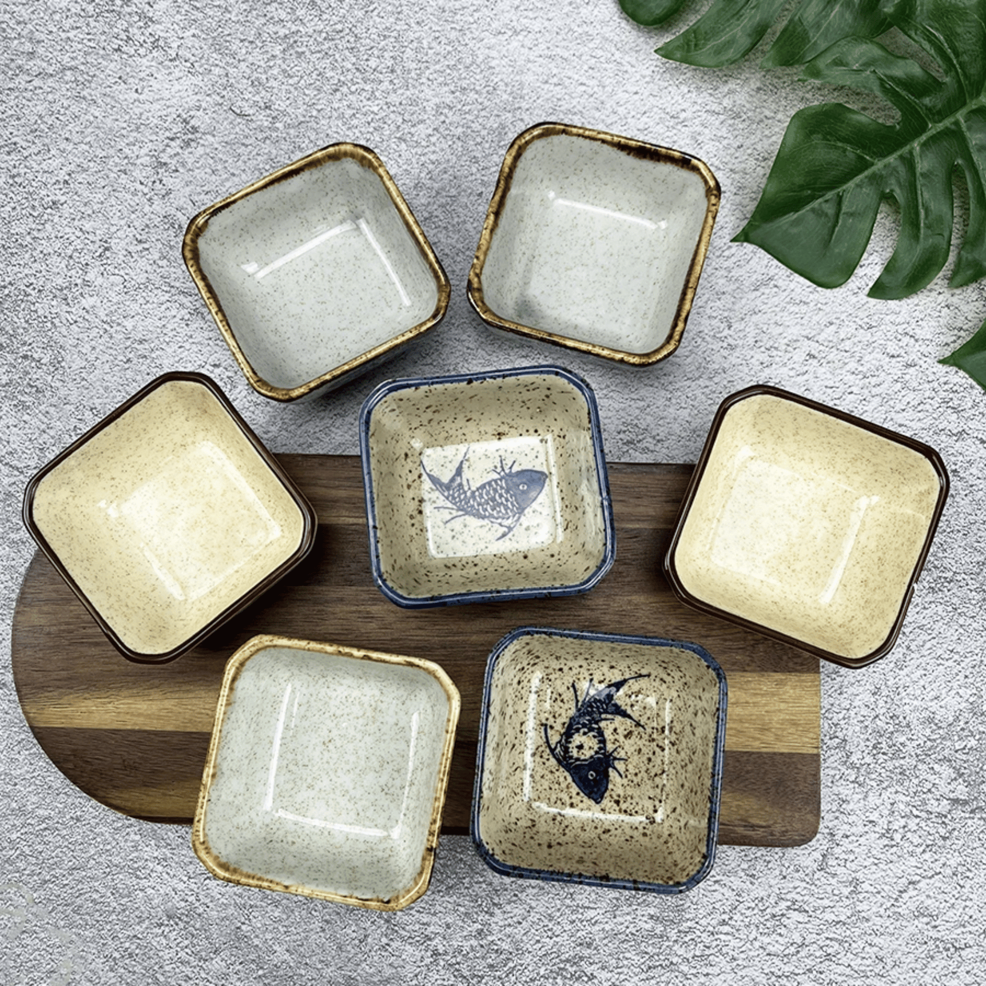 Set of square ceramic dishes with fish design on a wooden board, on a textured surface with leaves.