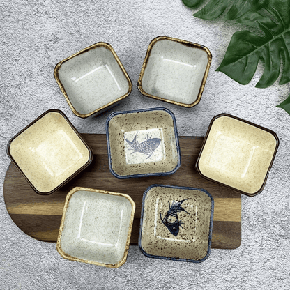 Set of square ceramic dishes with fish design on a wooden board, on a textured surface with leaves.