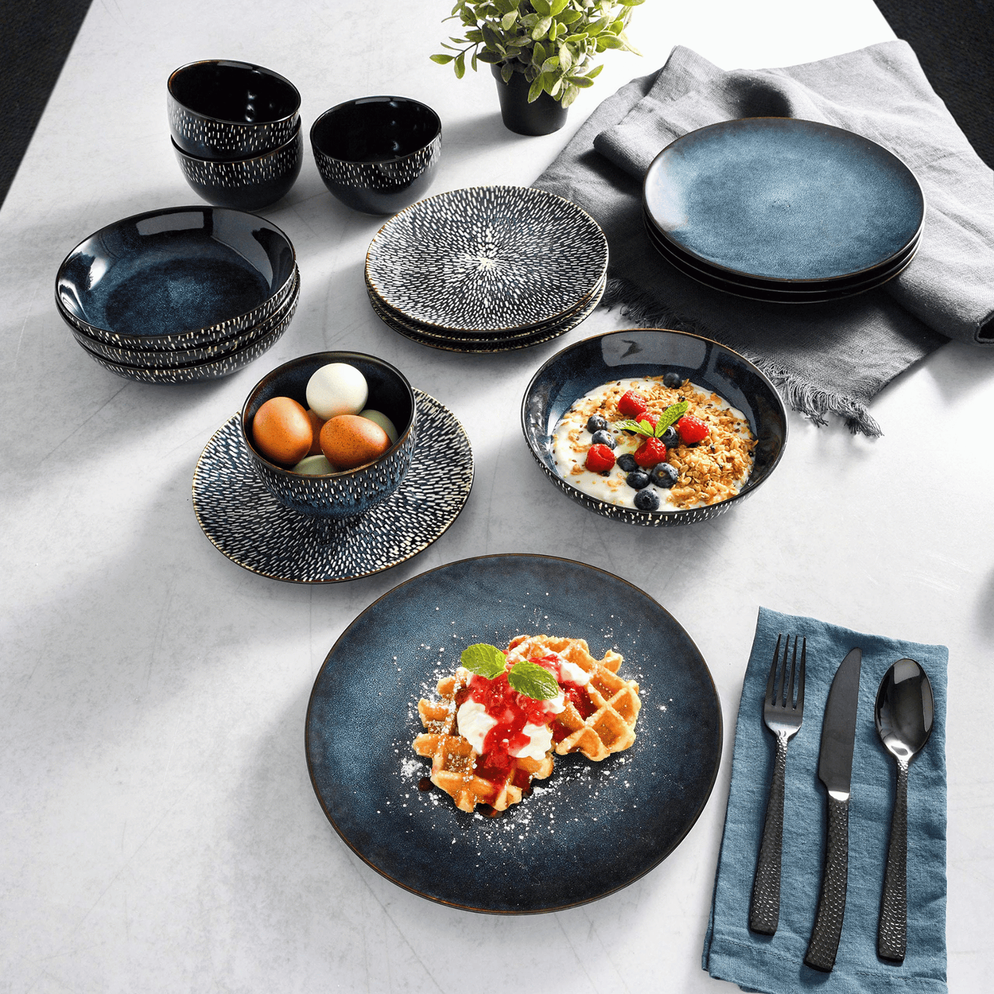 Set of black ceramic plates and bowls on a white tablecloth with food and cutlery.