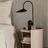 Modern black floor lamp on a round wooden side table next to a bed with beige bedding.