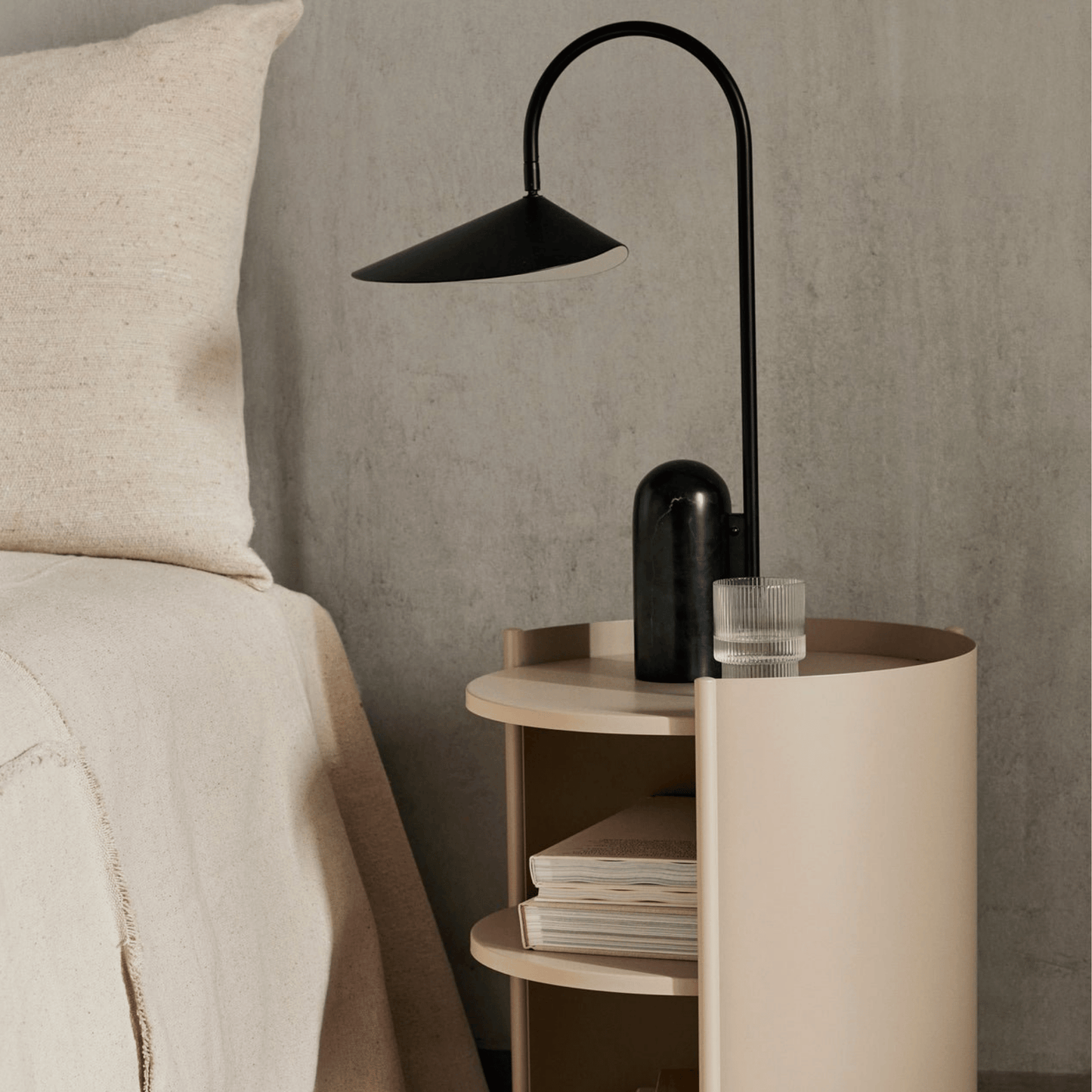 Modern black floor lamp on a round wooden side table next to a bed with beige bedding.