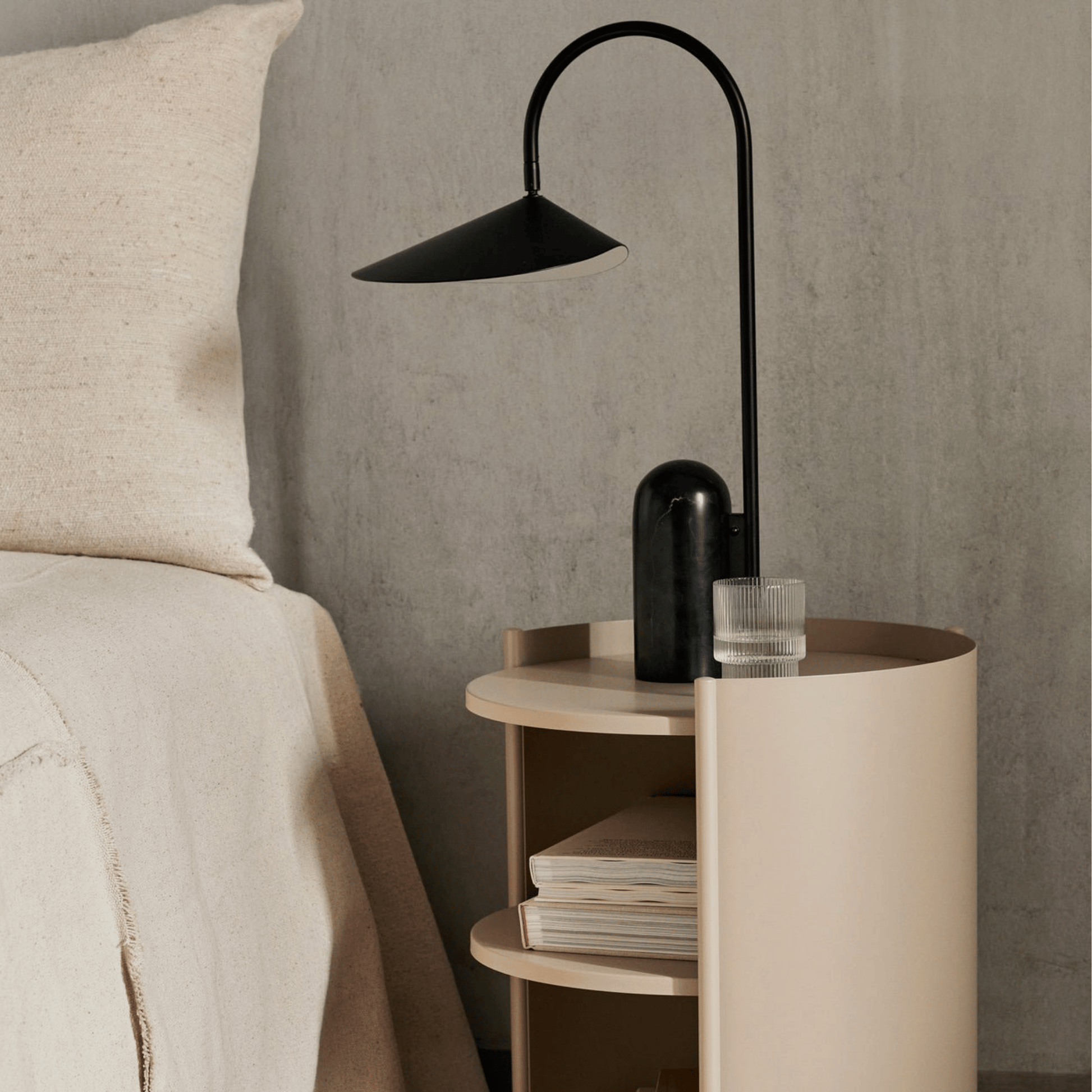 Modern black floor lamp on a round wooden side table next to a bed with beige bedding.