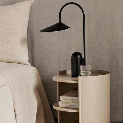 Modern black floor lamp on a round wooden side table next to a bed with beige bedding.