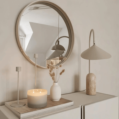 Round mirror on a wall with a lamp, candle, and vase on a shelf below.