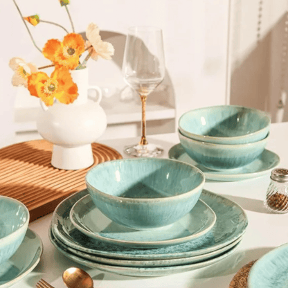 Set of blue ceramic dinnerware on a table with a vase of flowers and a glass.