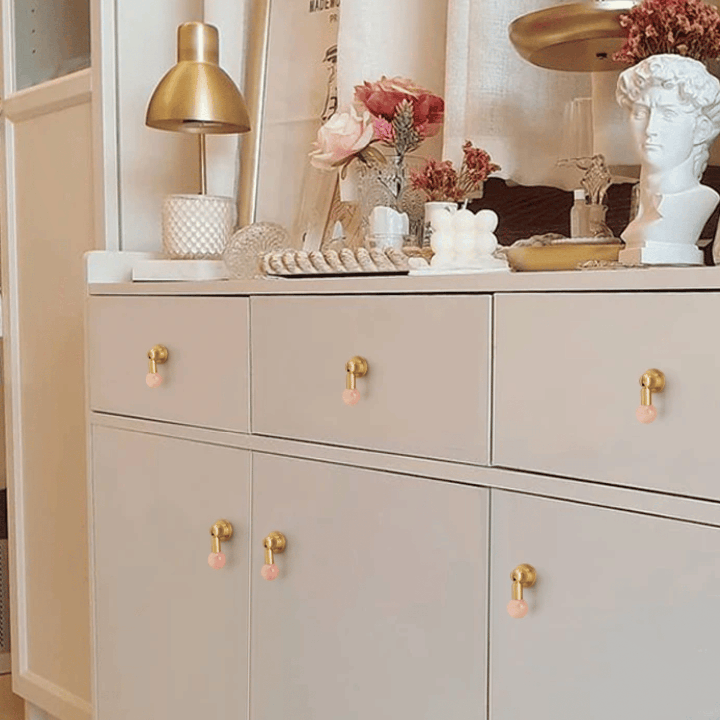 Beige cabinet with gold and pink knobs, decorated with flowers and a lamp.