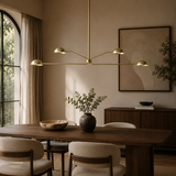 A modern brass multi-arm chandelier hangs over a wooden dining table in a warm, elegantly styled dining room with beige walls, upholstered chairs, and a large framed abstract print.