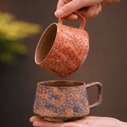 Two ceramic mugs with a speckled glaze held by hands against a brown background
